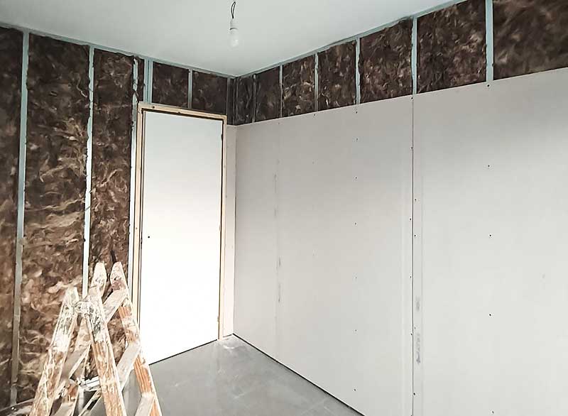 GS - Serveis Instalación pladur Habitación con una pared cubierta de aislamiento y una puerta visible en el fondo.